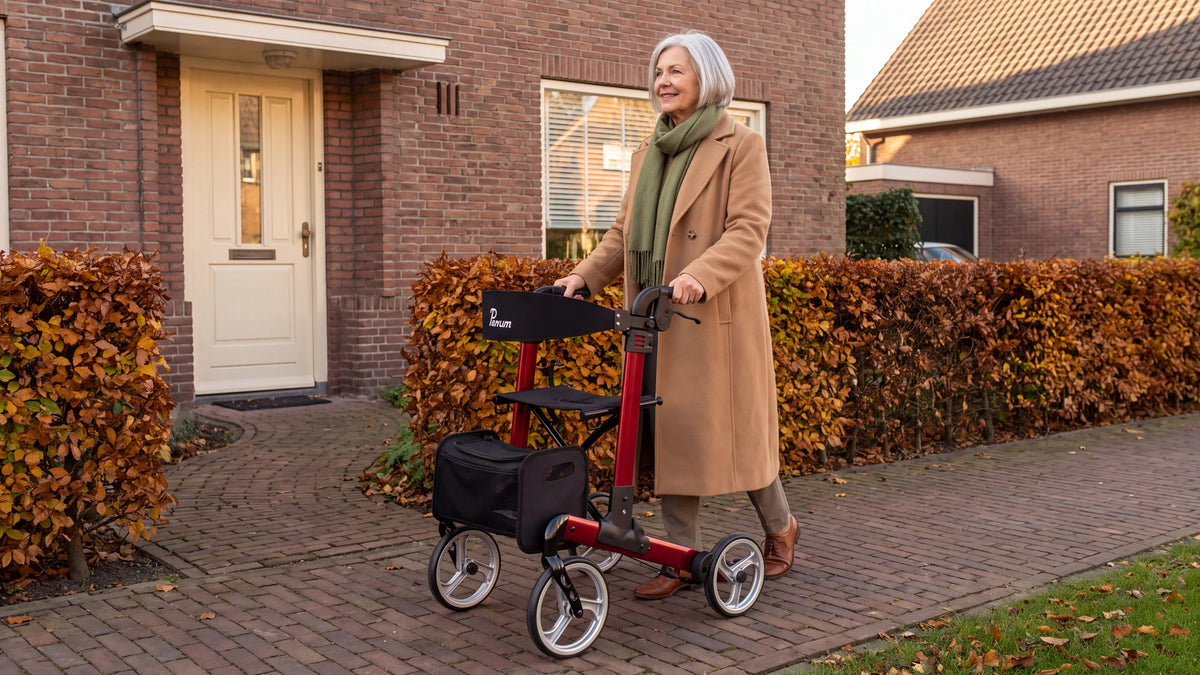 Parvum Luxo in ruby red; a woman in a camel coat steps out of the front door with the rollator, past a beech hedge.