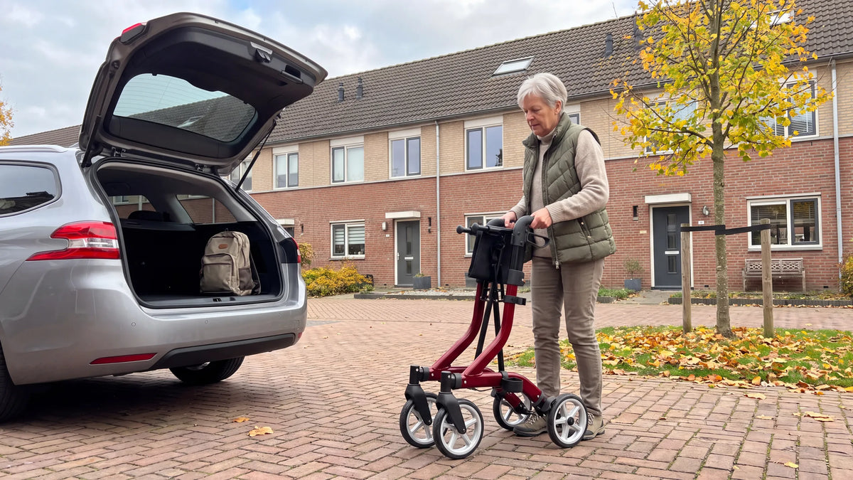 Ingeklapte Parvum SuperLight Plus in rood naast de open kofferbak; een vrouw pakt hem bij de handvatten.
